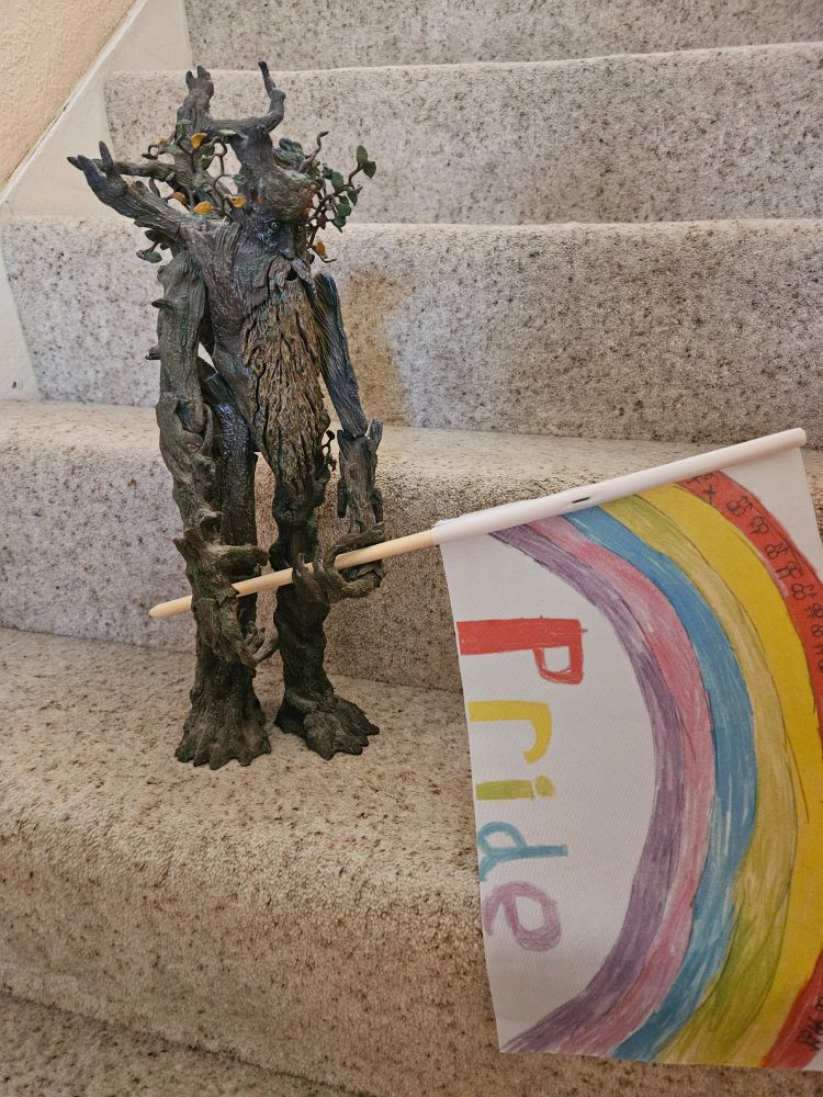 My beige carpeted stairs. A large action figure of Treebeard, a large tree like creature "Ent" from The Hobbit, holds a wooden sticked small flag in his long woody hands. The white flag shows a child's picture of a rainbow with rainbow text underneath reading: Pride.