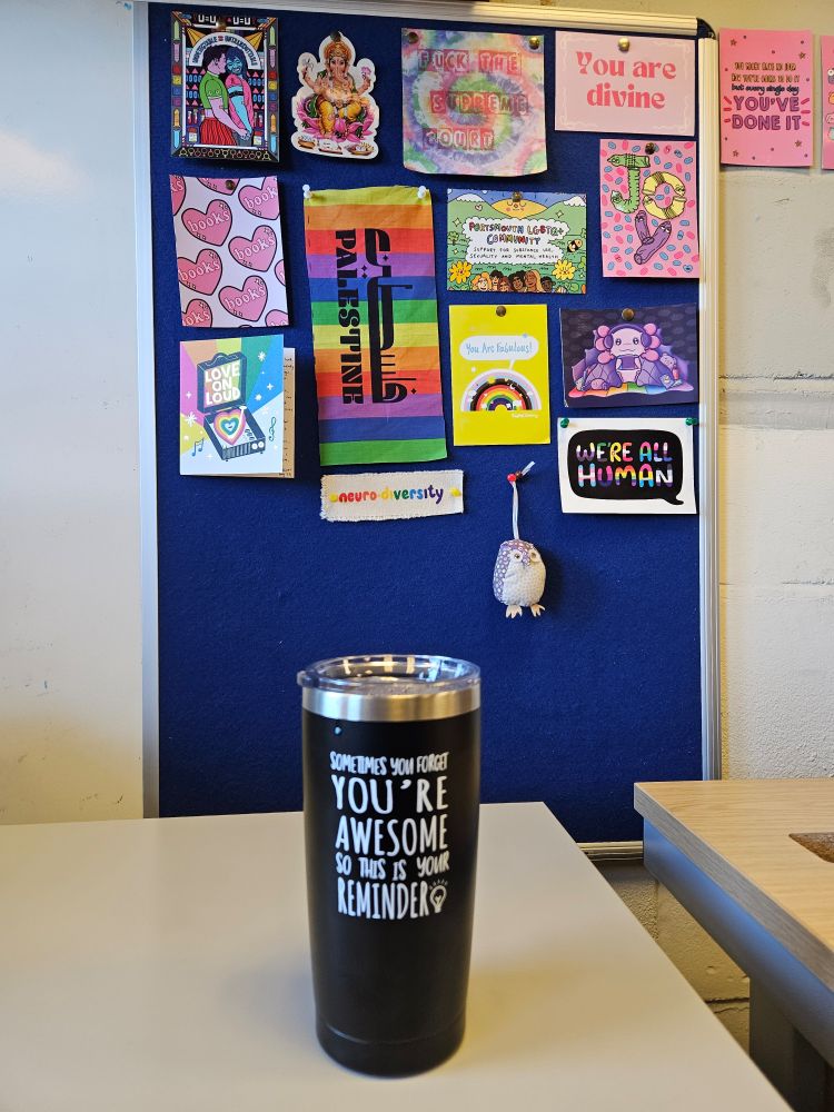 A coffee thermos with the above phrase on. It sits in front of my board, slowly filling with very colourful queer postcards, lots of rainbows, love hearts, ganesh and condoms