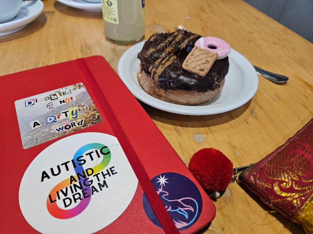 A light brown coffee table, my bright red diary sits next to a delightful chocolate topped cronut. my diary has three stickers, Autistic and Living the Dream logo, Trans Aware Cancer Care logo and a sticker by Artie which reads "Disabled is not a dirty word".