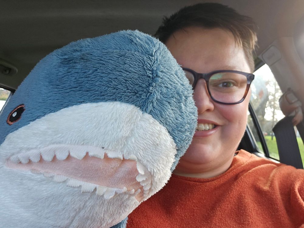 Me, a large white person seen from the shoulders up. I've got scruffy hair and my glasses are slightly steamed up (argh the weather!). Im holding up a blue grey blahaj/shark from IKEA which I will be waving manically court side]