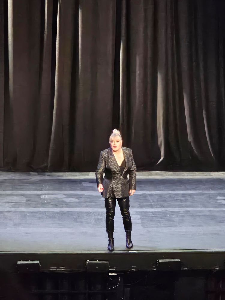 Suzy, a tall, blond haired woman on stage. She is wearing red lipstick, red nails, a plain black jacket, and plain black leather trousers. The stage is sceneless and propless. Black curtains hang at the upstage.
