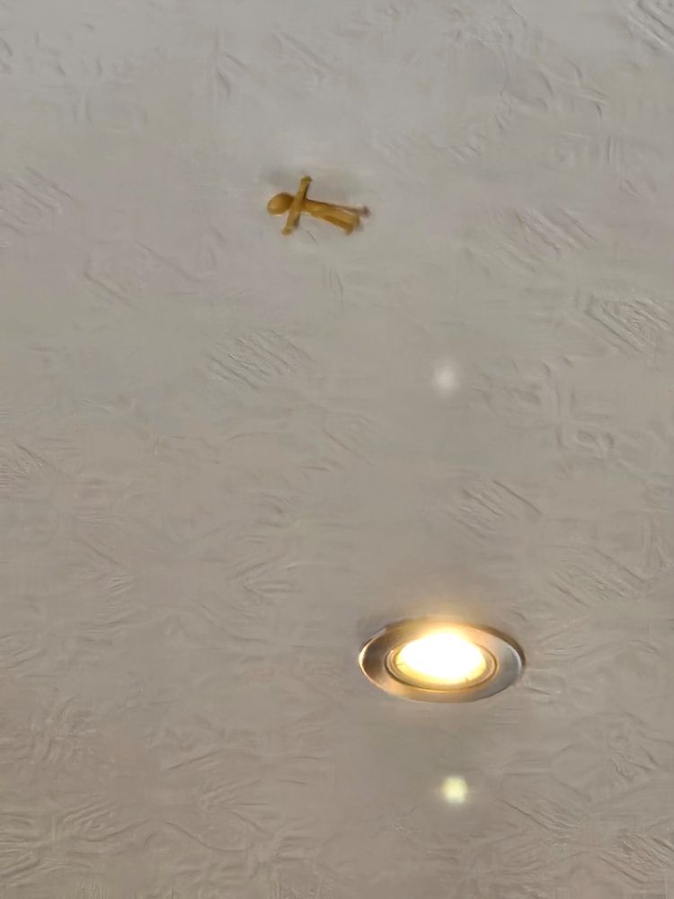 A little stretchy yellow person stuck to a white textured ceiling, an arm and a leg hang loose
