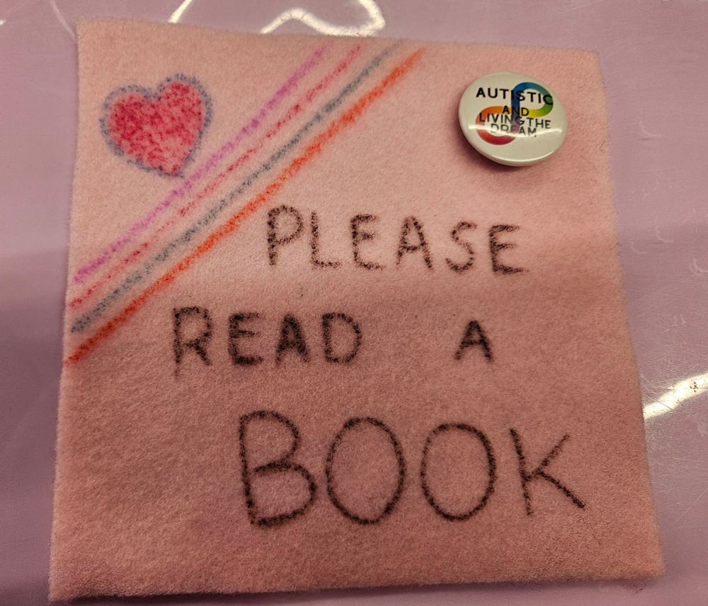 A pink felt square I decorated today. Top left is an felt tip pink heart with a blue outline. Four lines on an angle are underneath in pink, red, blue and orange. Large black handwriting at the centre of the square says: please read a book