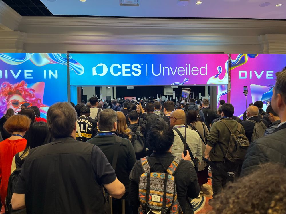 Crowd of people entering a room with screens around the door reading “CES | Unveiled”  and “DIVE IN”