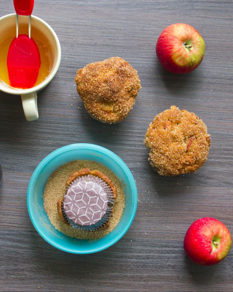 zwei Muffins, die in Apfel-Zimt-Mischung getunkt wurden, daneben Äpfel, eine Tasse mit geschmolzener Butter und eine Schüssel mit Zimt-Zucker, in der kopfüber ein Muffin steht