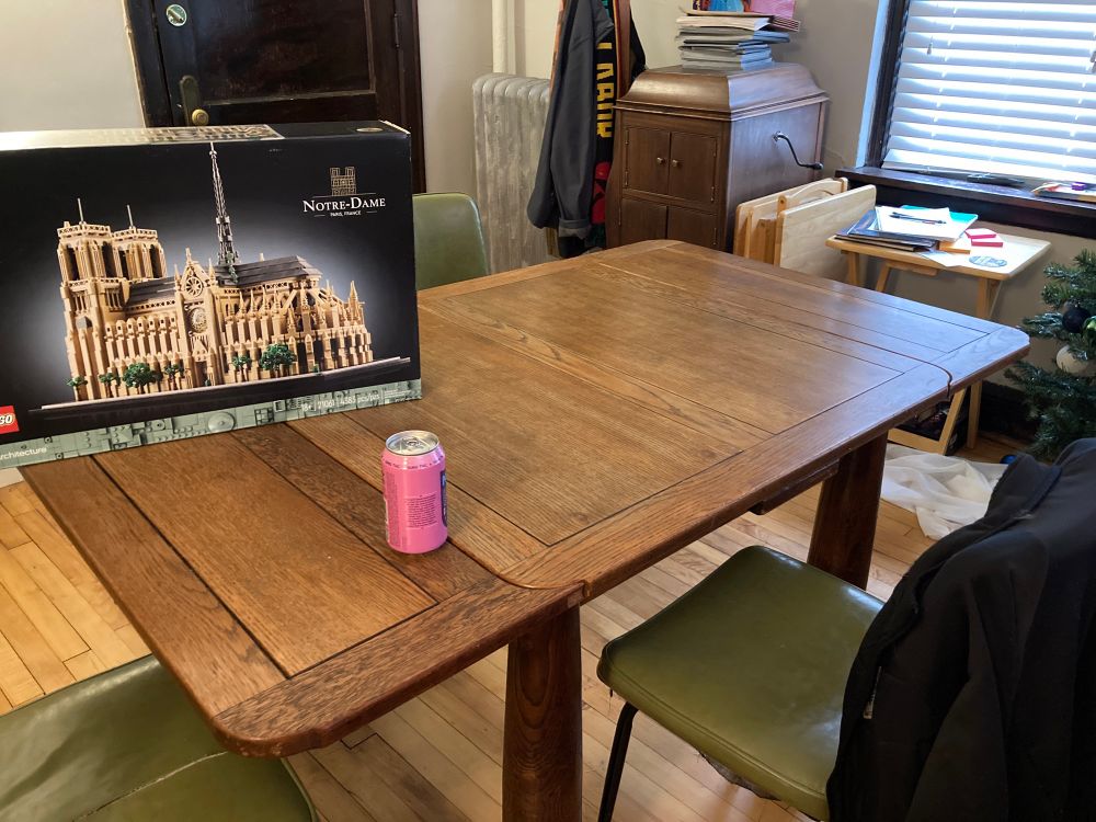 A large, boxed Lego set of Notre Dame sits on a large table in a room. There is also a beverage on the table. 