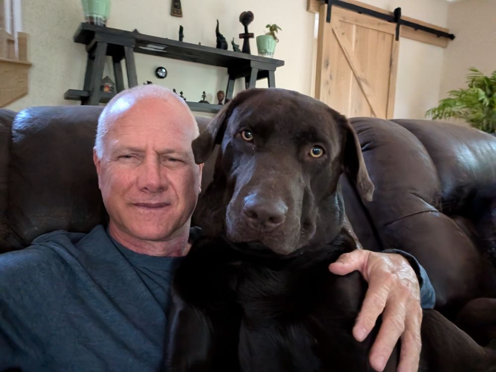 A white man with very short gray hair sitting in a recliner with his hand around a chocolate Lab sitting on him. Both are looking at the camera.