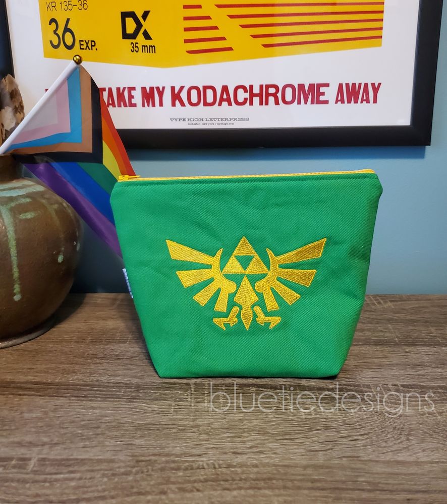 A green zip bag with the hyrule crest in yellow siting on a surface with a rainbow flag in the background.