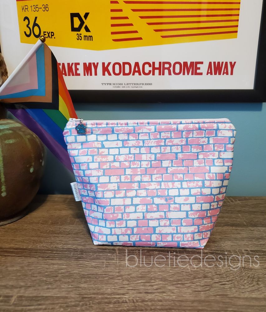 A pink, white, and blue brick patterned zip bag sitting on a surface with a rainbow flag in the background.