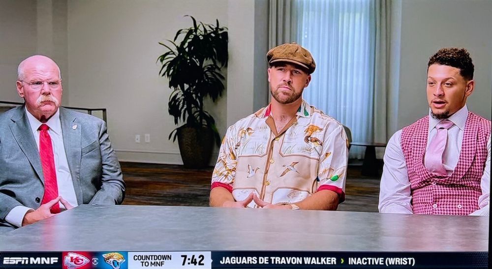 Photo of a Kansas City Chiefs press conference with Andy Reid, Travis Kelce, and Patrick Mahomes. Reid is wearing a gray suit and red tie with a lapel pin. Kelce is wearing a camp-style short-sleeved shirt with a bird print, along with a  suede cap, and Mahomes is wearing a white collared shirt, a pink checkered double-breasted vest, and a pale pink tie.