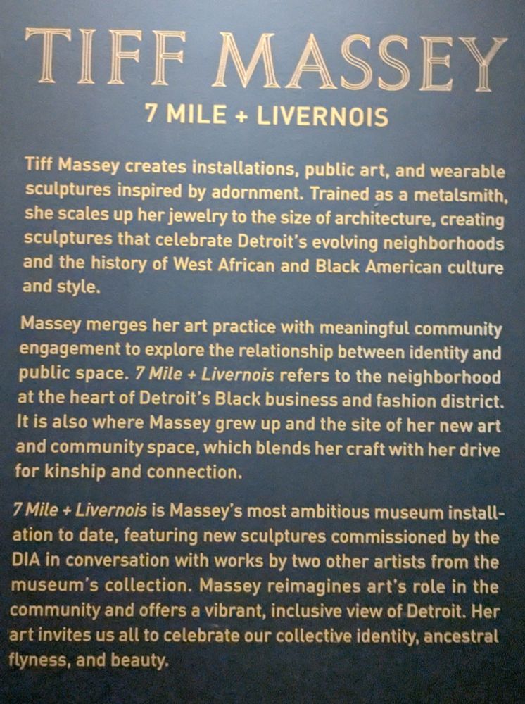 The image shows an informational plaque about artist Tiff Massey and her exhibition titled "7 Mile + Livernois". Here's the complete text:

TIFF MASSEY
7 MILE + LIVERNOIS

Tiff Massey creates installations, public art, and wearable sculptures inspired by adornment. Trained as a metalsmith, she scales up her jewelry to the size of architecture, creating sculptures that celebrate Detroit's evolving neighborhoods and the history of West African and Black American culture and style.

Massey merges her art practice with meaningful community engagement to explore the relationship between identity and public space. 7 Mile + Livernois refers to the neighborhood at the heart of Detroit's Black business and fashion district. It is also where Massey grew up and the site of her new art and community space, which blends her craft with her drive for kinship and connection.

7 Mile + Livernois is Massey's most ambitious museum installation to date, featuring new sculptures commissioned by the DIA in conversation with works by two other artists from the museum's collection. Massey reimagines art's role in the community and offers a vibrant, inclusive view of Detroit. Her art invites us all to celebrate our collective identity, ancestral flyness, and beauty.