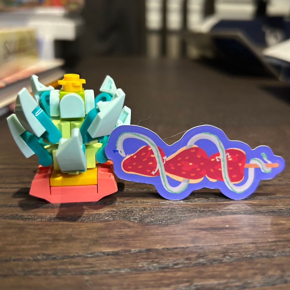 A lego plant that appears to be a succulent of some sort. It has spiky bits around a central small yellow flower. 

Next to it is a sticker of a tiny dragon wrapped around a stick of candied strawberries. 

Today’s hint for the sticker:
Noodleji’s favourite sweet treat at the fair.