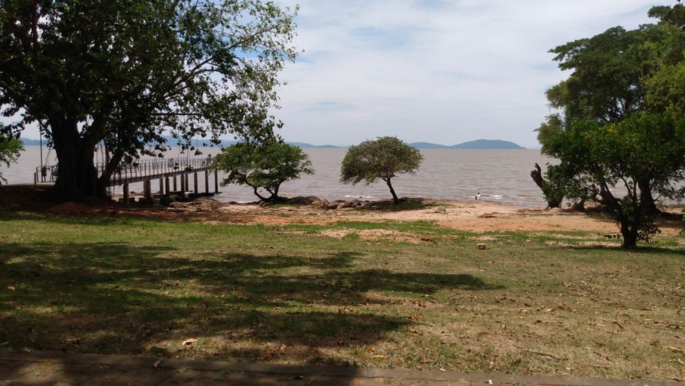 Vista da praia da alegria, Guaíba RS 

Uma praia com árvores e pedras, um pier e morros ao longe depois do corpo d'água.