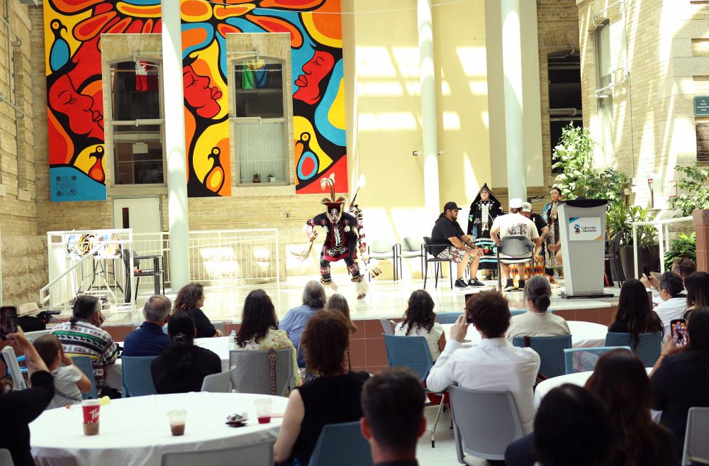 Traditional Indigenous dancer on stage with circle of drummers as people watch in Brodie Atrium 