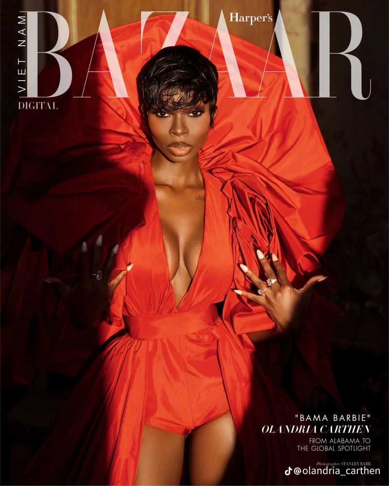 A Black woman stands confidently in a dramatic red gown with oversized, structured sleeves fanning around her shoulders. She poses against a warm, softly lit indoor backdrop, wearing a deep-plunge bodice, glossy makeup, and long nude nails and a “wet look” pixie cut. The Harper’s Bazaar Vietnam Digital masthead overlays the image.