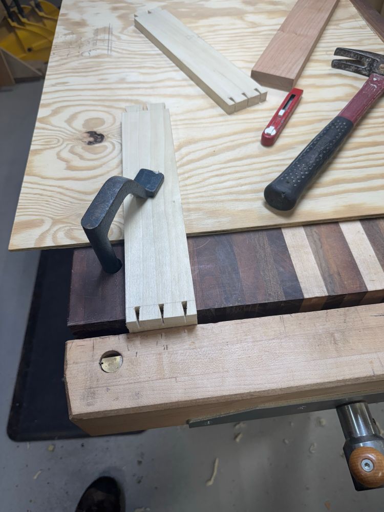 Dovetailed board being held on a woodworking bench by an iron holdfast. Also in shot are another board, a red marking knife and a hammer. There’s a big piece of plywood on the bench starting about six inches from the end.