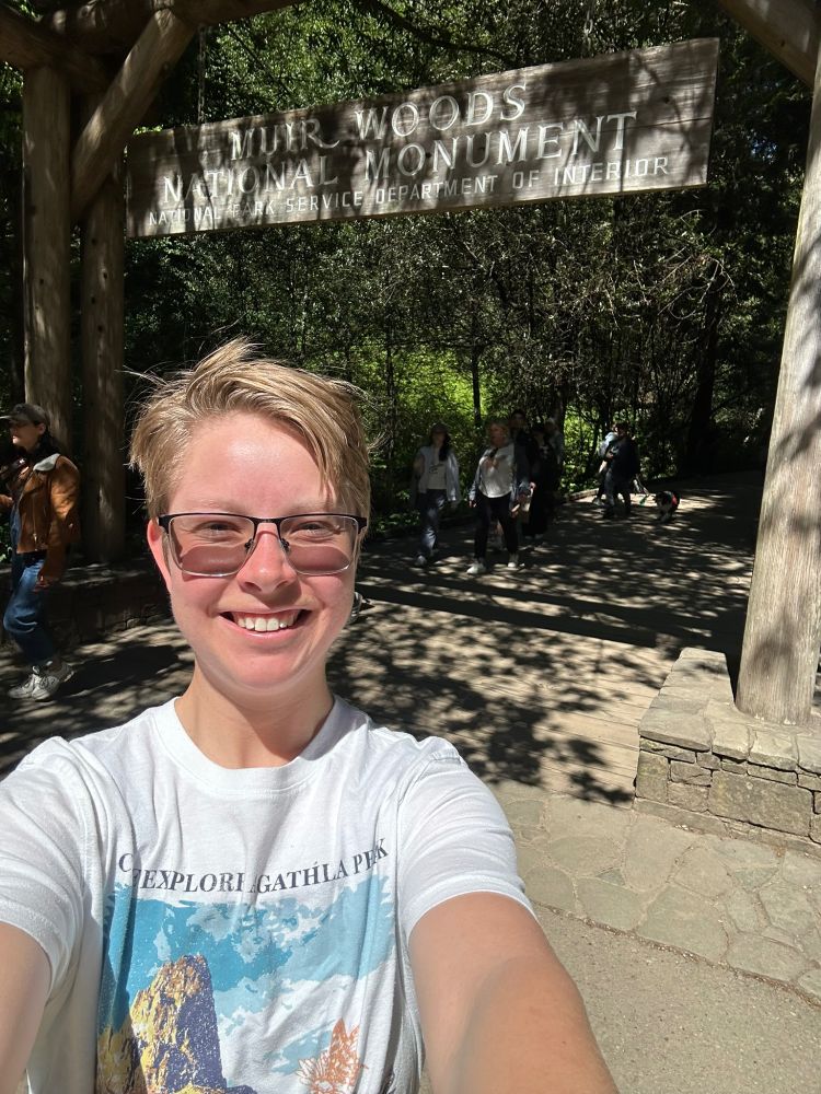 A photograph of myself infront of the "Muir Woods National Monument" sign! I am white with blond hair and glasses, squinting into the sun and the camera 