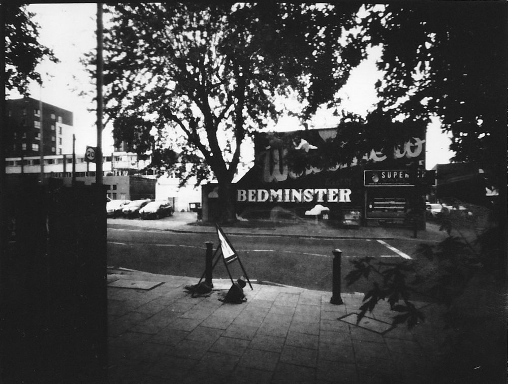 Black and white pinhole photograph of a road in front of a wall with street art reading 'Welcome to Bedminster'