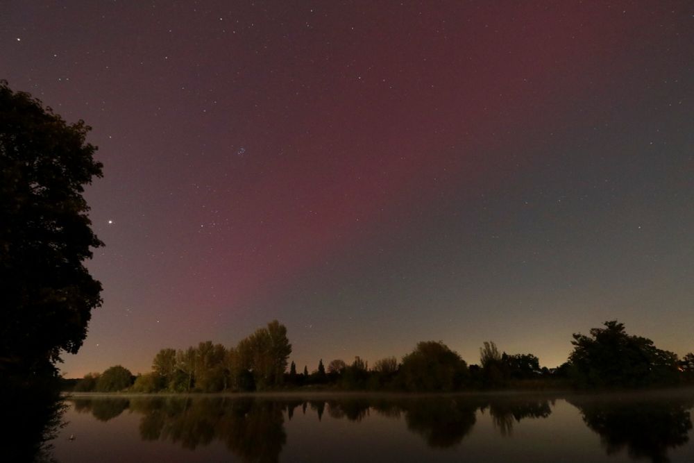A starry sky with pink northern lights above a river surrounded by trees. There's a light fog on the surface of the water, which reflects the trees like a mirror.