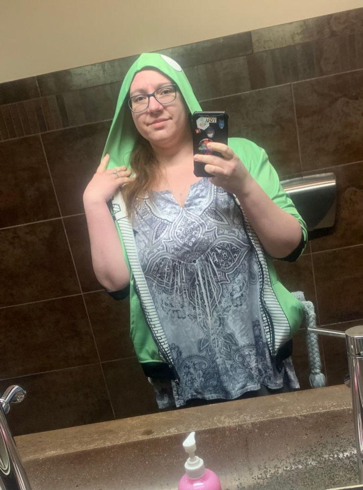 Woman in a green Gir hoodie and grey and white print shirt 
She is taking a selfie in a bathroom mirror 