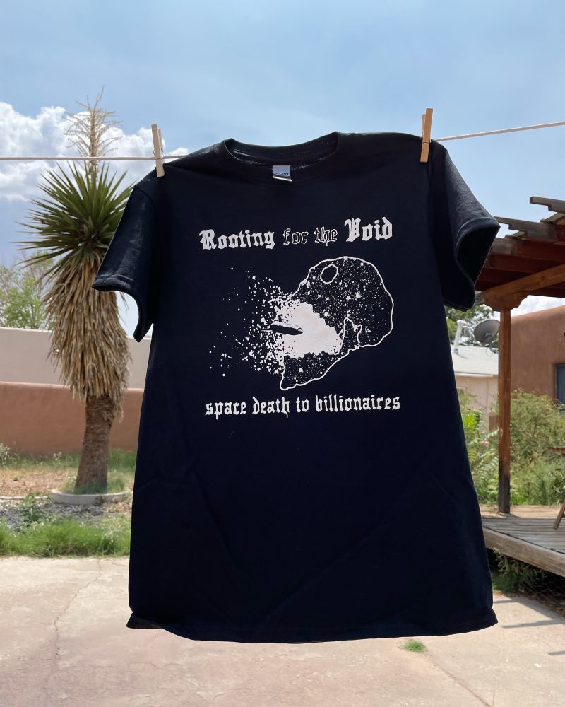 A black shirt hangs from a clothesline with a white print showing a spaceship being lost in a skull shaped cosmos. The text “Rooting for the Void” runs across the top and “space death to billionaires” under the image. 