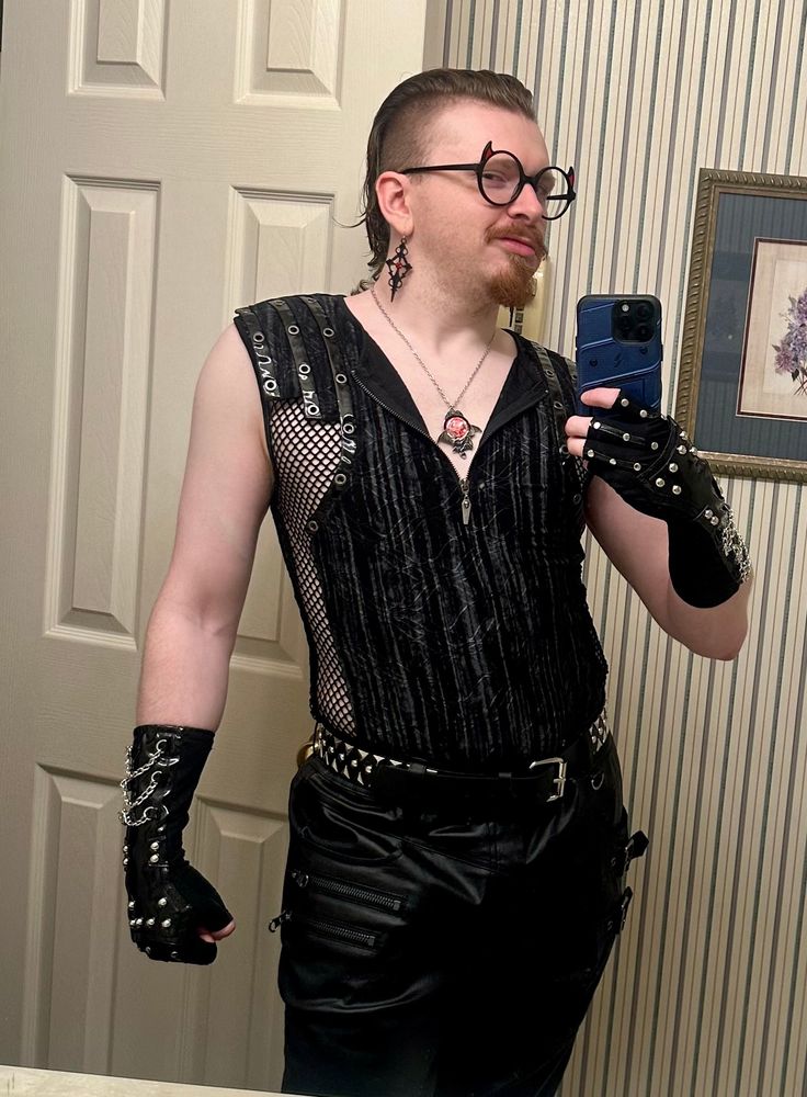 Me, a lean white he/they homo twunk with a strawberry blonde/auburn mohawk mullet hybrid that’s been slicked back and sprayed down, and a matching goatee and mustache. I’m wearing a snazzy black sleeveless top with an unzipped collar, vertical grey striped dye pattern, mesh pits and sides, and vertical leather strips with metal eyelet rings on the shoulders. My pants are black leather with zippers and buckles below the pockets, the waist secured by a triangular + circular studded black leather belt. I have on studded and chained fingerless black leather gloves that run halfway up my forearms, and black diamond shaped dangly earrings with a pointy cross inside and a red gemstone in the center. My glasses frames are large, black, and round with red devil horns on the rims, and there is a silver chain around my neck with a silver bat amulet housing a round blood red gem to symbolize the blood moon.