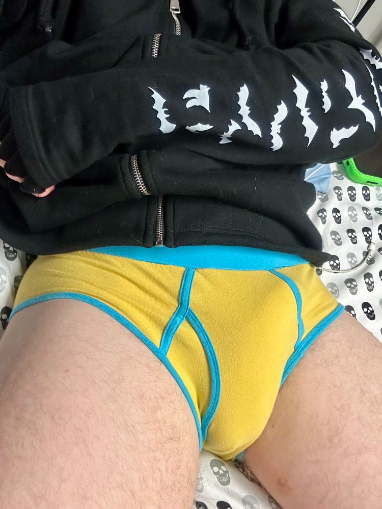 Me, a cute goth he/they homo boy in a black hoodie with white bats down the sleeve and a pair of bright yellow ringer briefs with light blue elastic. 