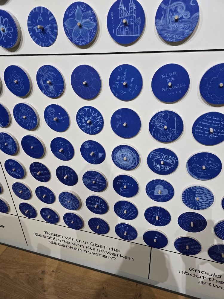 A white wall covered with blue circles that people have drawn on with white pencils. There are various pictures and doodles. One says FREE PALESTINE with a Palestinian flag. Others have hearts, faces, flowers etc 