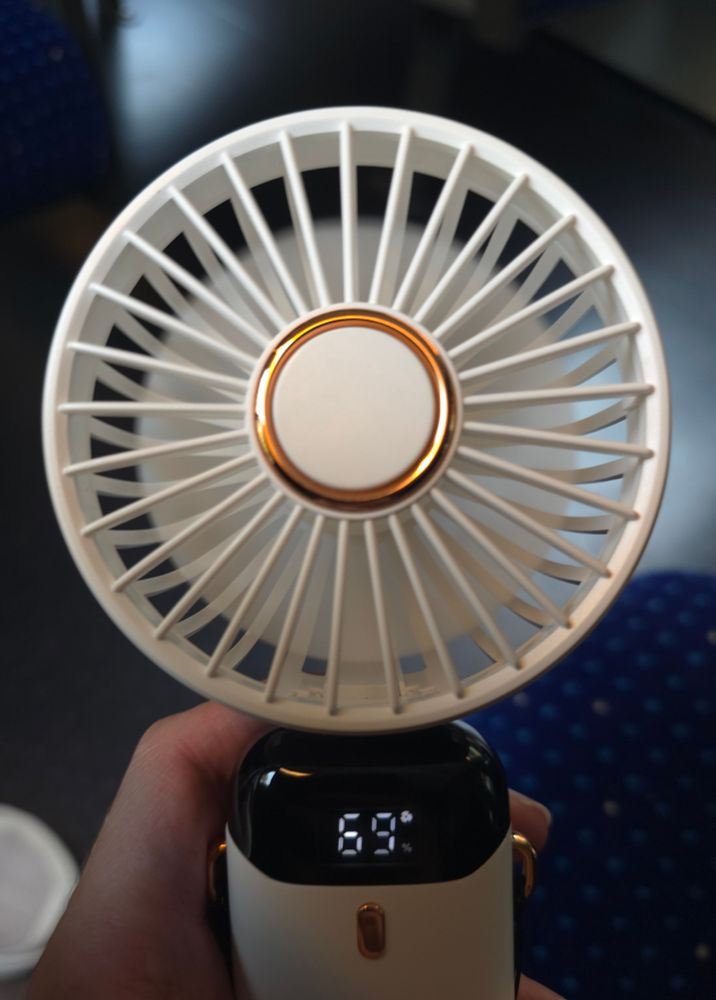 A white electric handheld fan with a gold ring around the centre of the fan and a black display showing the battery read out that says 69%. Held by a white hand with a blue train seat in the background 
