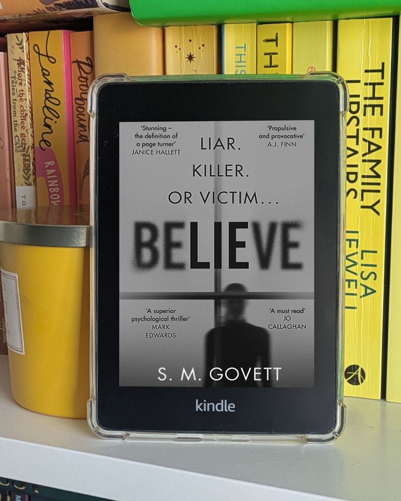 Photo of a Kindle Paperwhite in front of a bunch of orange and yellow books and a yellow candle jar on a shelf. The kindle screen shows the front cover of 'Believe' by S.M. Govett. 