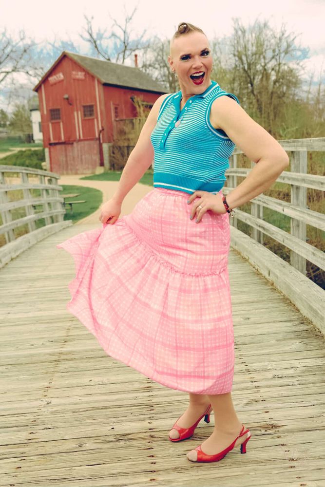A woman in a pink ankle length skirt and blue sweater vest is pictured mid-spin on a wooden bridge with an historic mill in the background. She is smiling and wearing red lipstick. Her shoes are red kitten heels. Her nails are blue shimmer with pink glitter