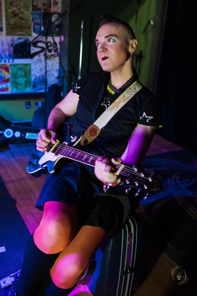 A woman sits on a suitcase kick drum playing the guitar. There is a bar scene behind her. The lighting is poor but getting better