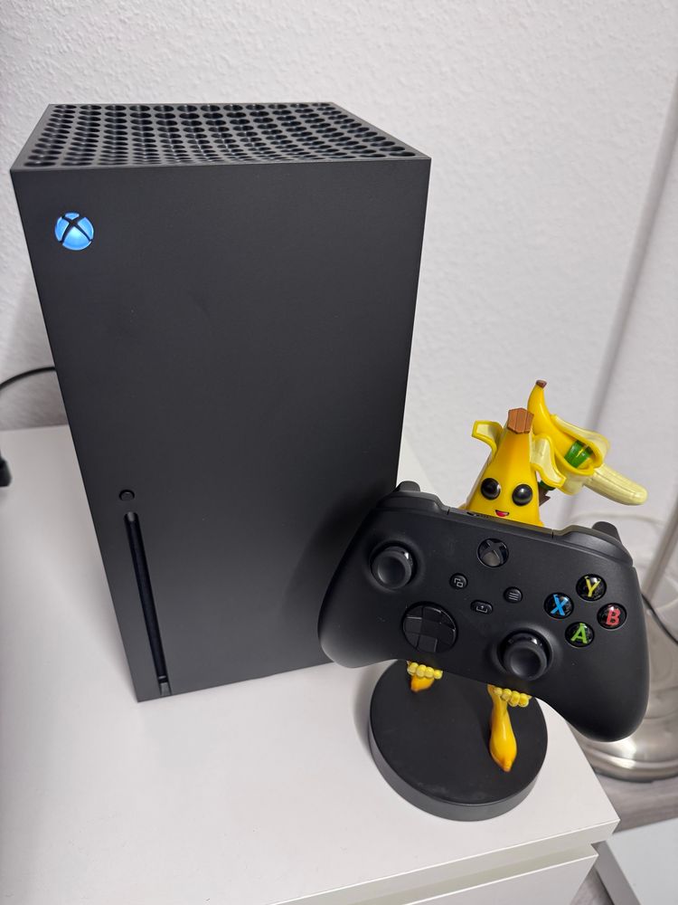Eine schwarze Xbox Series X auf einem weißen Schrank, rechts daneben ein schwarzer Controller auf einem Fortnite-Controller-Halter