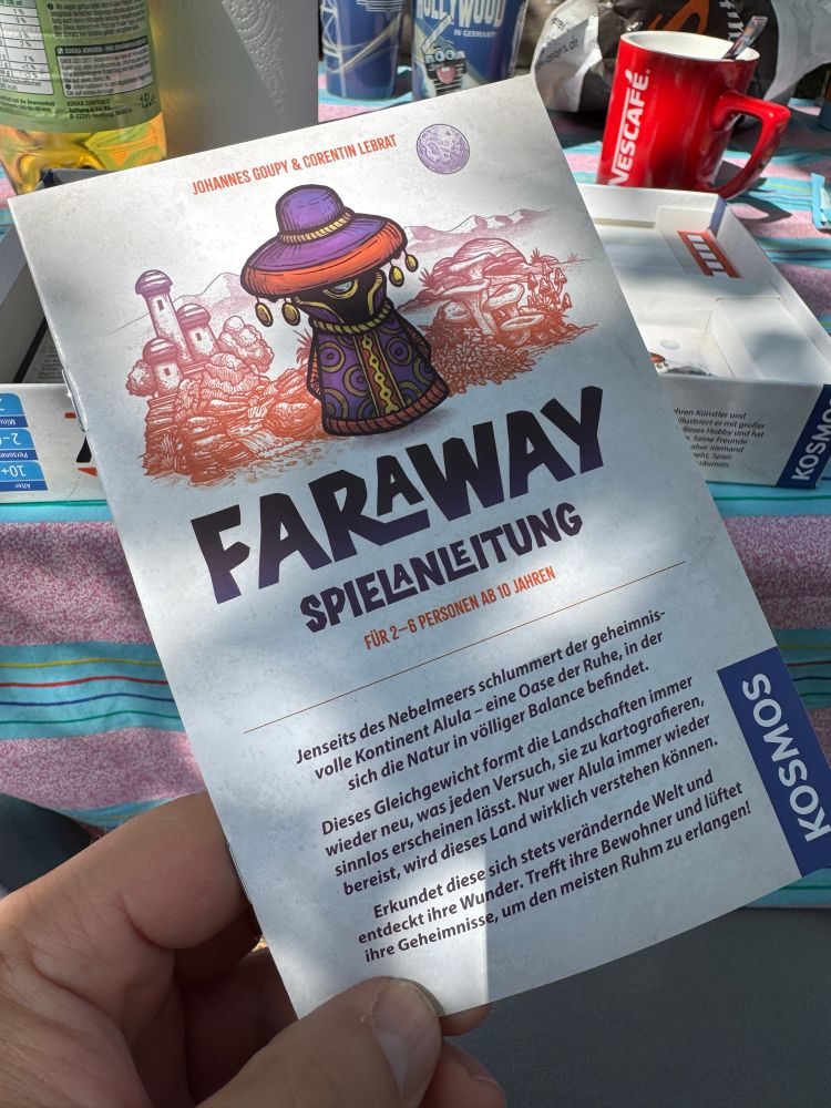 Eine Hand hält die Spielanleitung von „Faraway“. Im Hintergrund sieht man einen Tisch, auf dem der Rest vom Spiel liegt, sowie eine Kaffeetasse und diverse Becher. 
