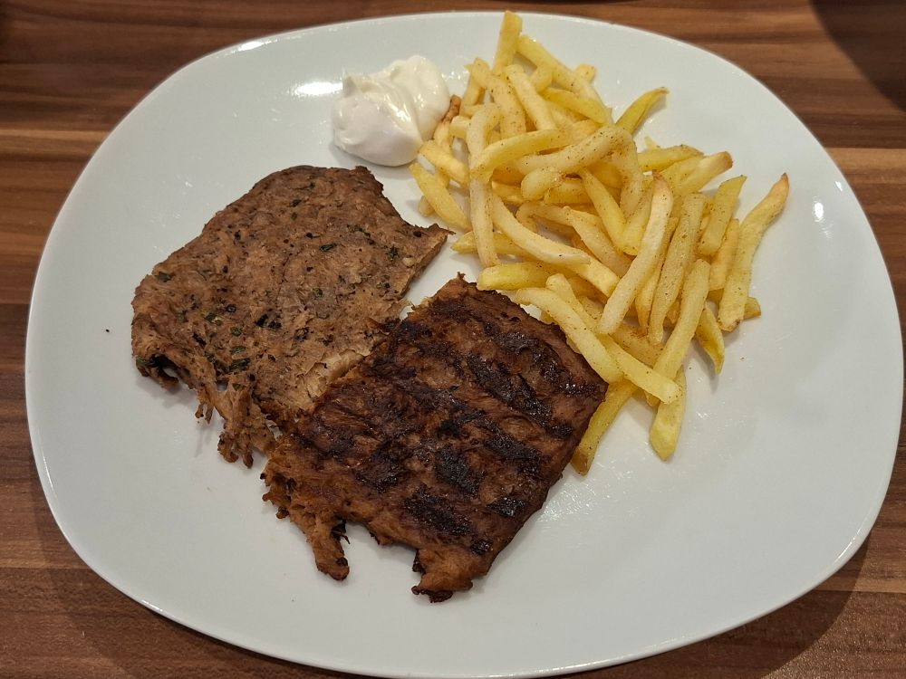 Ein weißer Teller und auf dem Teller zwei unterschiedliche Soja-Bigsteaks und Pommes mit einem Klecks Mayo.