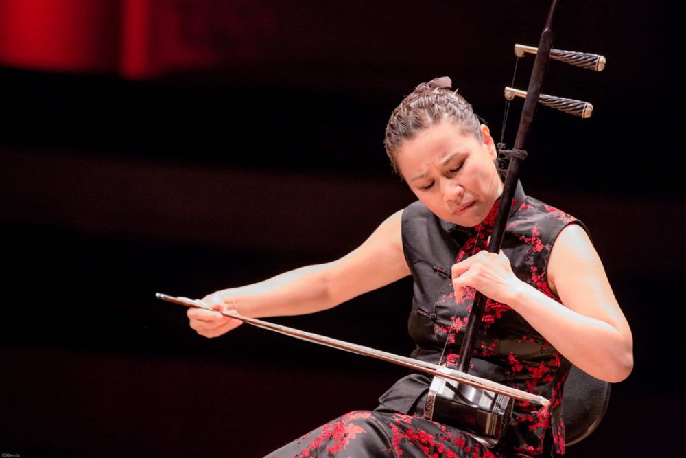 A photo of an Asian lady playing a two stringed instrument with a bow. It looks like a hexagon cylinder with a stick on it and two large cones at the top of the stick for tuning.