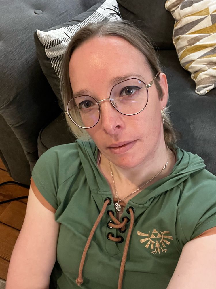 Jacky wearing a Legend of Zelda inspired dress looking straight in the camera with a slight smile. The dress is green and has copper accents, it’s got a v-neck collar with laces and a green hood.