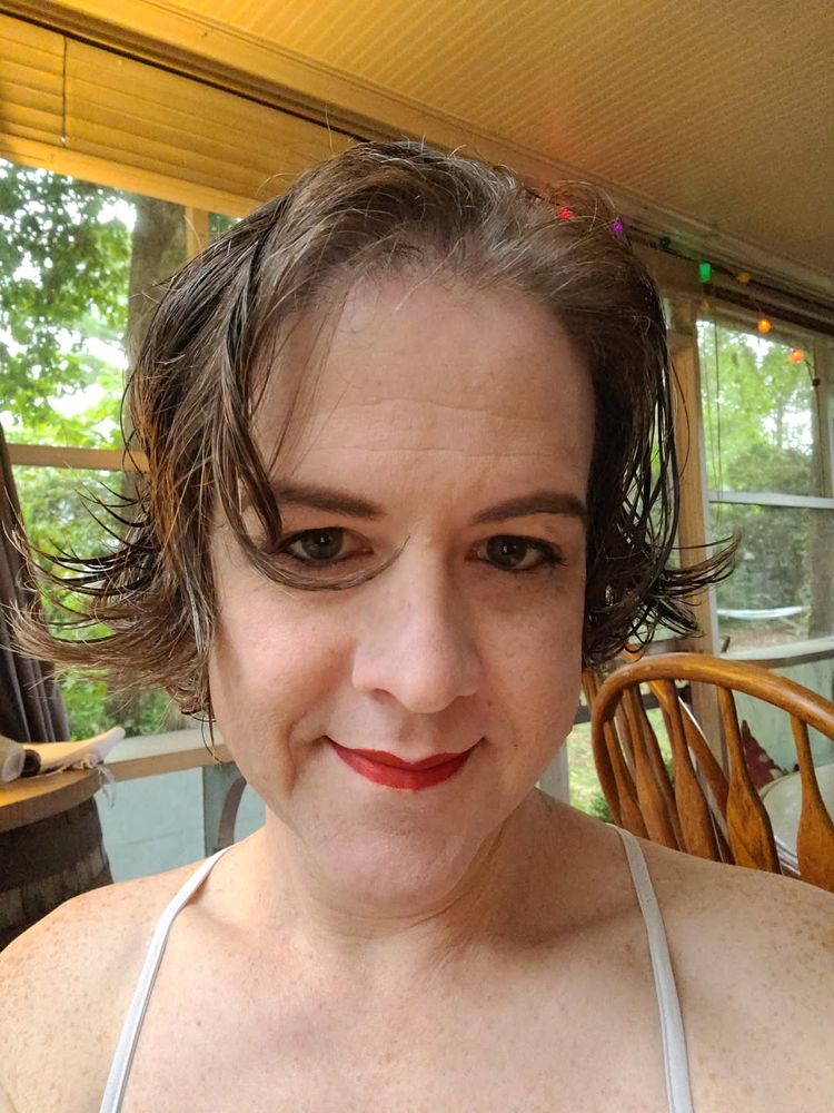 Face picture of white woman shoulders up with makeup done while wearing a spaghetti strap dress, red lips, curly hair that's wet and flaring out presently