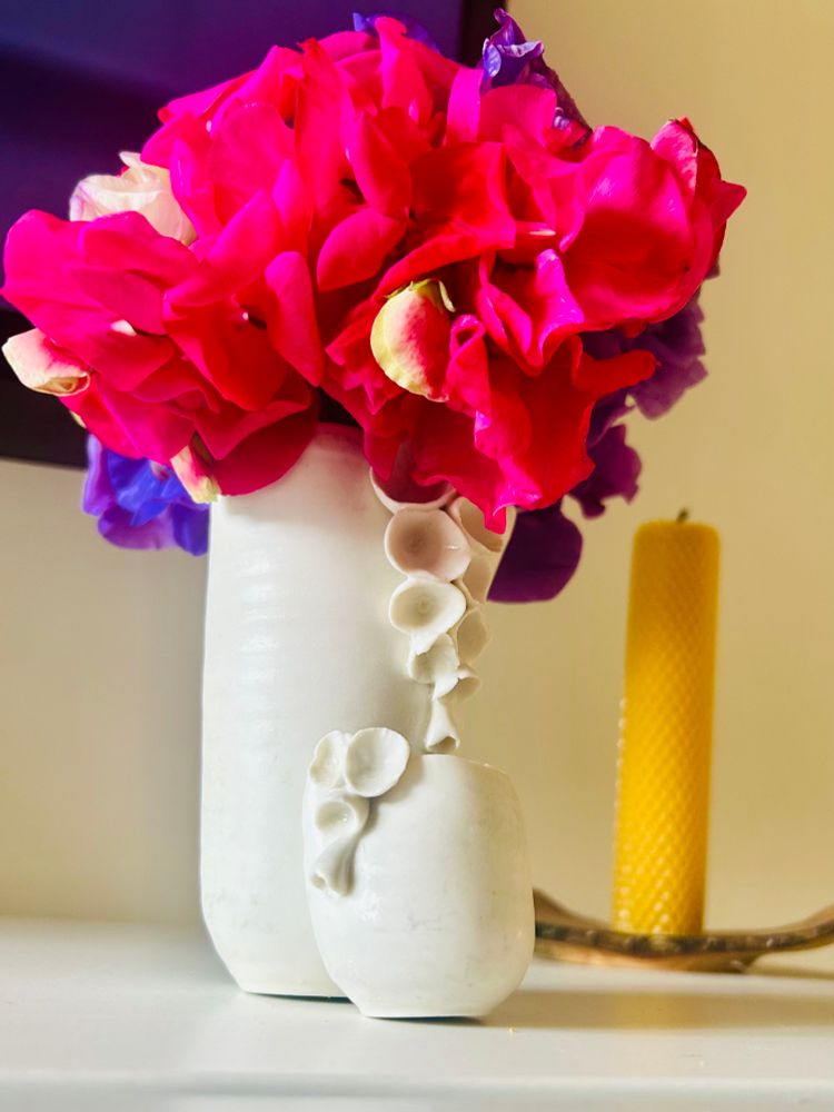 A bright, bold bouquet of pink, magenta, and purple sweet peas bursts from a tall, white ceramic vase adorned with delicate flower-like details. A smaller matching vase sits in front, echoing the same sculptural motif. Behind them, a tall honeycomb beeswax candle in a brass holder adds a soft golden touch. The scene is warm, textured, and quietly elegant — a perfect balance of colour and calm.