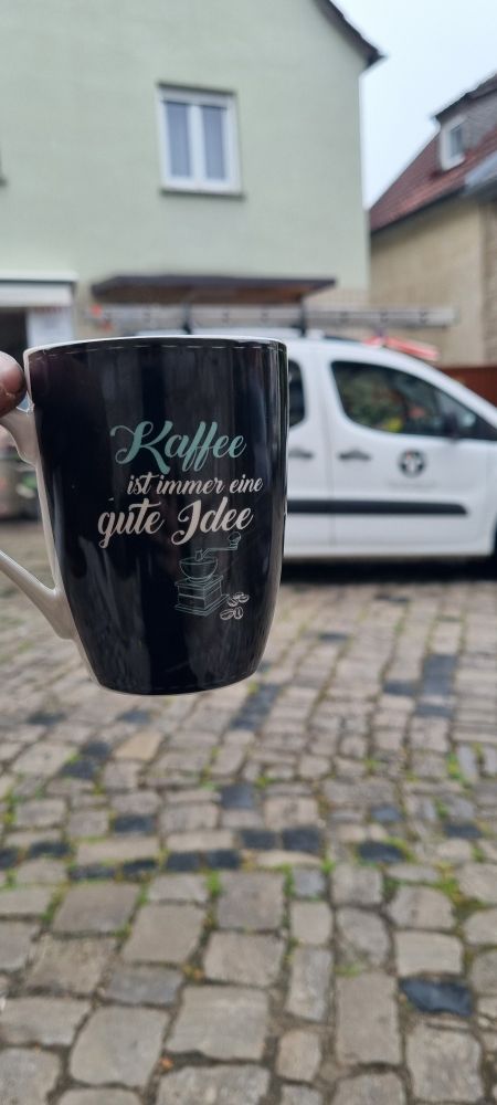 Mittig links im Bild eine schwarze Kaffeetasse, Beschriftung "Kaffee ist immer eine gute Idee". Im Hintergrund ein weißer Kastenwagen mit Kaminkehrer Logo auf der Seite. Der Boden ist altes Kopfsteinpflaster. Hinter dem Auto ist ein grünes Haus zu sehen mit Schaufenster.