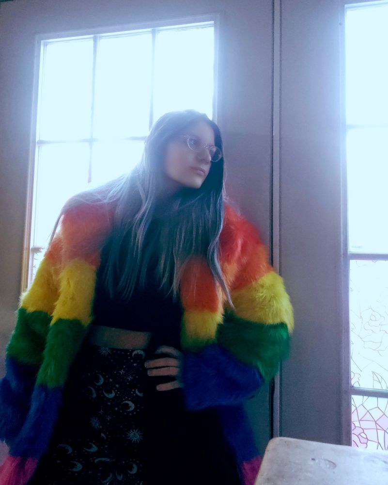 Woman with glasses and black and grey hair standing with hand on hip. She's leaning on French doors and looking out the windows. She's wearing a rainbow coat made of faux fur, a black crop top, and black tights with white moons and suns printed on them. 