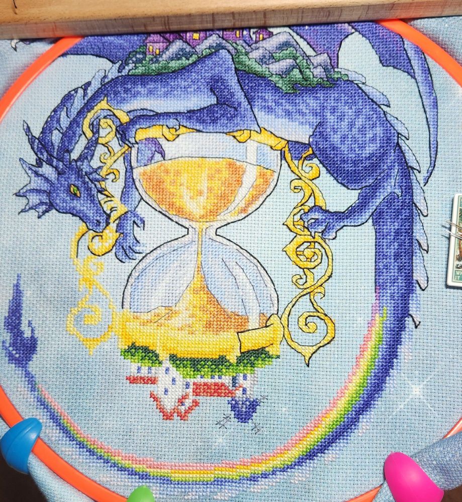 A cross stitched piece of a large blue and purple dragon wrapped around a golden hourglass, with about 75% of the black back stitching finished
