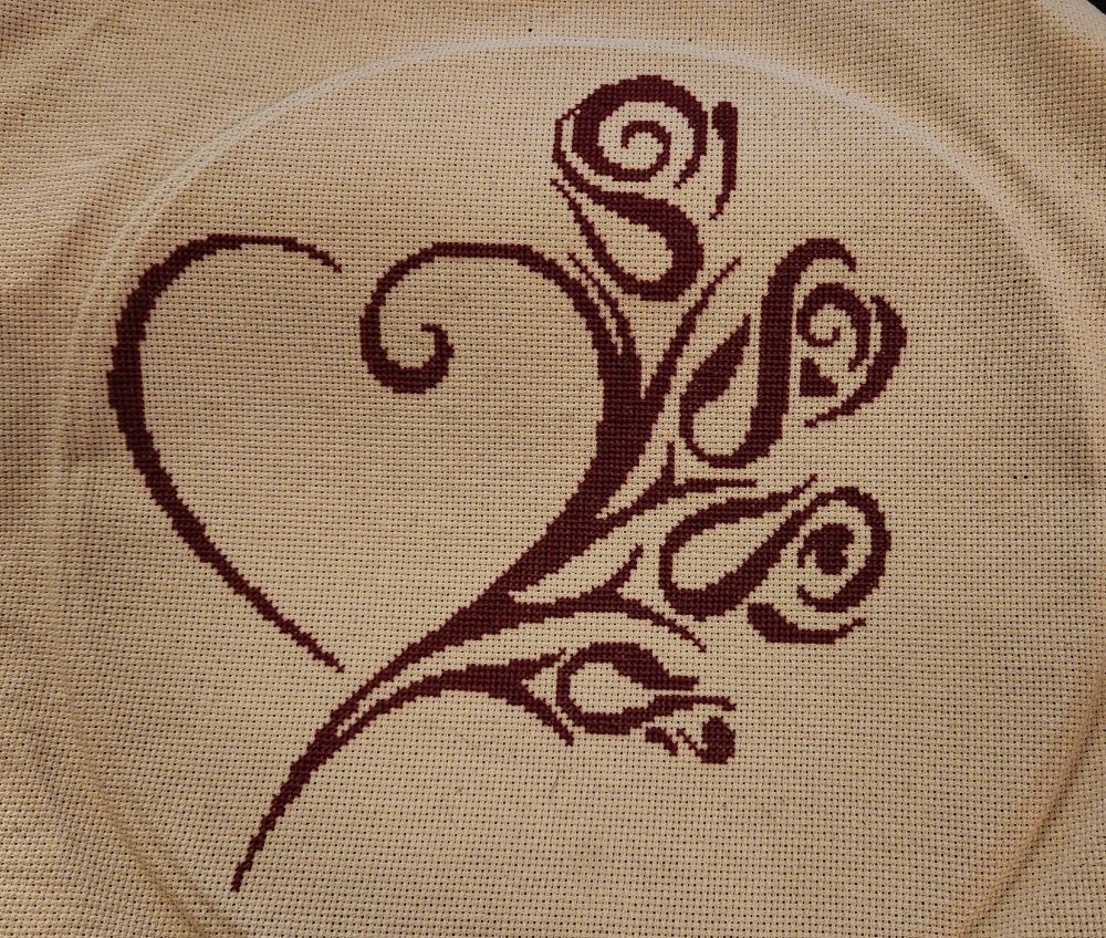 A finished cross stitch worked in a wine colored embroidery floss on an antique white aida. A large heart with stylized roses and hearts to the right coming off the side of the heart. The fabric still bears the impression of the embroidery hoop around the outside of the finished stitching.