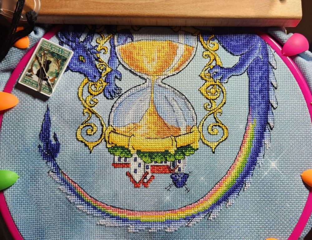 A mostly finished cross stitch of a big purple dragon wrapped around a golden hourglass with a village on its back and a lighthouse type building on the bottom of the hourglass. Pattern is titled The Dragon That Keeps Time. The Dragon is missing black outlining around the tail and the bottom of the hourglass is mostly finished with black outlining stitches.