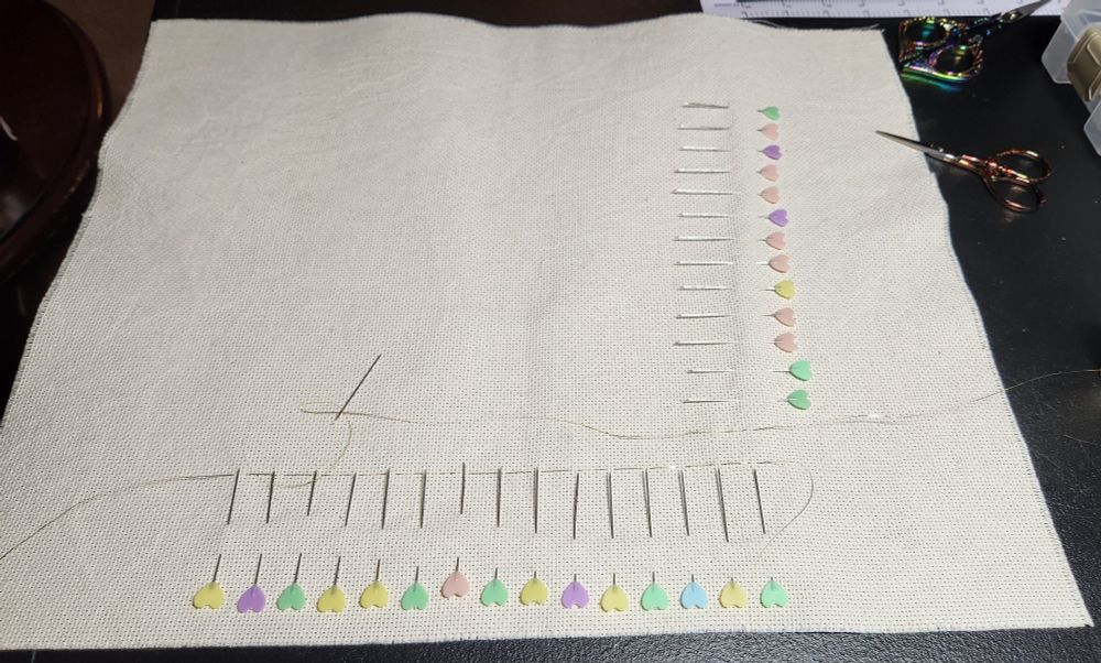 A piece of off-white aida fabric stuck with lots of straight pins around the edges, to make a 140x140 count grid broken up in blocks of 10. The straight pins have heart shaped heads in various pastel shades. There is a thin line of gold sulky thread across the bottom row of pins, the start of a cross stitching grid.