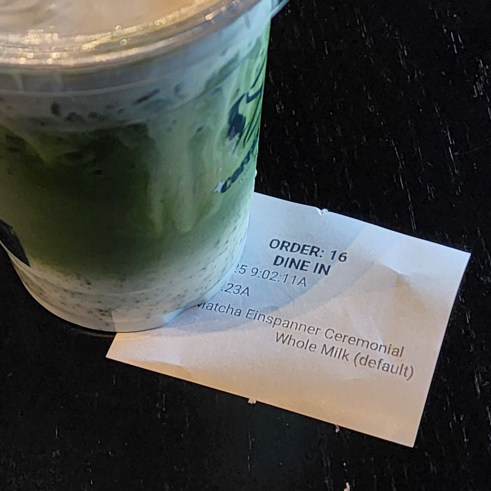 On a wooden table painted black, sits an order of matcha einspanner and the call receipt for the order.
