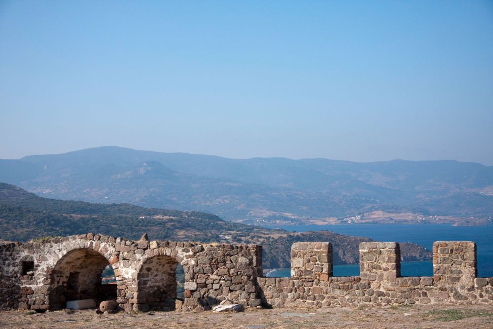 Lesbos:
Oude stenen muur met bogen op de voorgrond, uitkijkend over een prachtig landschap met heuvels en bergen in de verte. Achter de muur loopt de kustlijn met helderblauw water van een zee of groot meer. De lucht is helderblauw en er zijn geen wolken zichtbaar, wat duidt op mooi weer. De kleuren van het landschap zijn natuurlijk en het geheel straalt een rustige, historische sfeer uit.