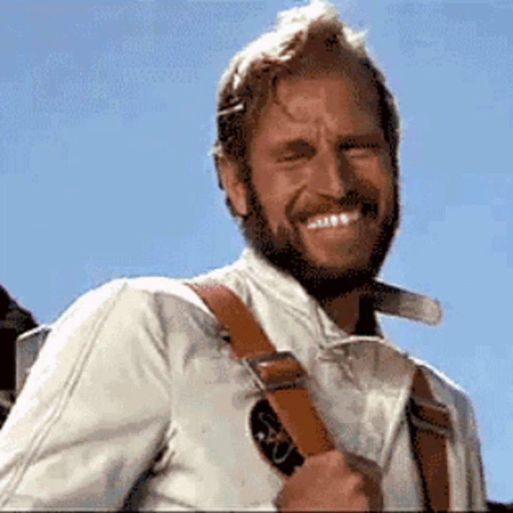 a man with a beard is smiling and wearing a white shirt and brown straps