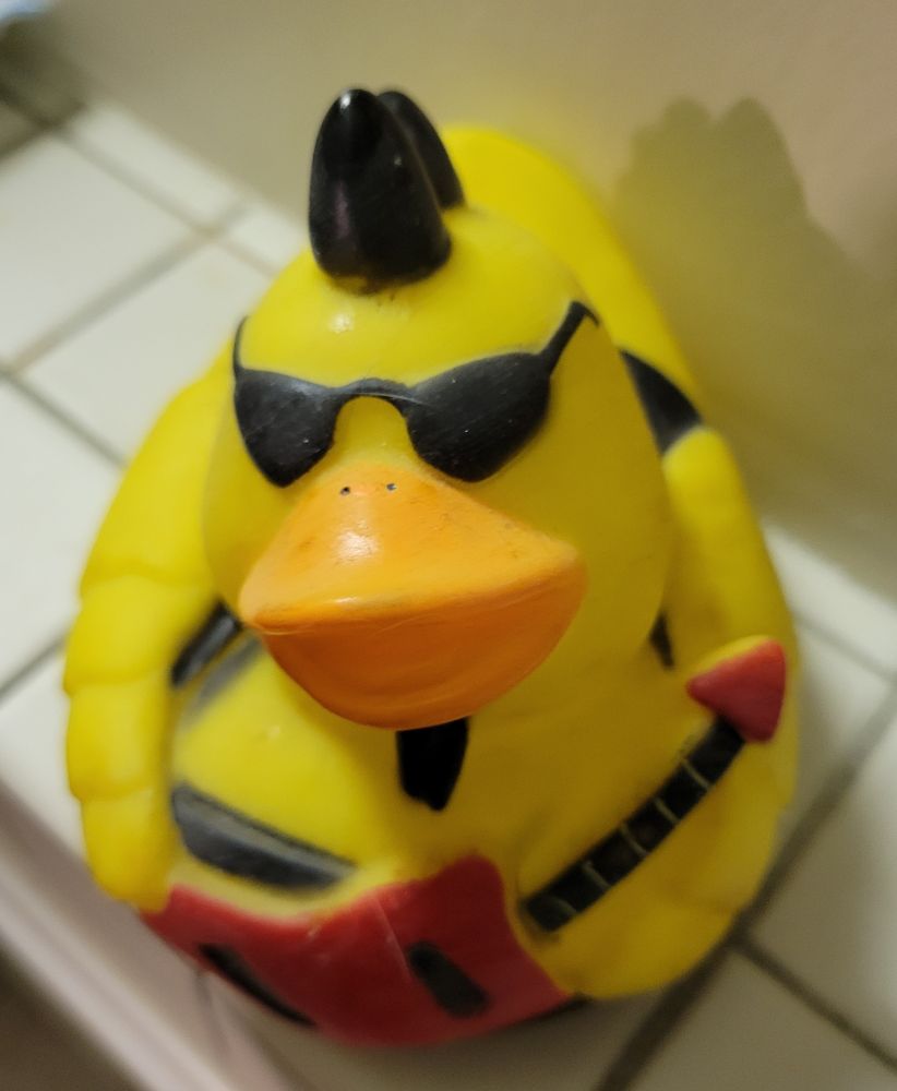 A yellow rubber duck with an orange beak sports a black spiked mohawk, black sunglasses, and a black soul patch.  They are styled to look like a rock star clad in black leather and wielding a bright red guitar.  Strong Drag King vibes.