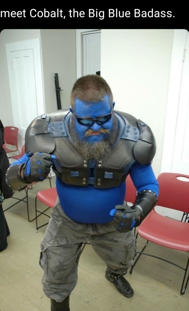 Dave participating in a superhero LARP dressed as his original character Cobalt, aka the Big Blue Badass.  Cobalt has short dark brown hair, a robust salt-and-pepper beard, vibrant blue skin, dressed in leather gear for (American) football and dark gray camo pants.